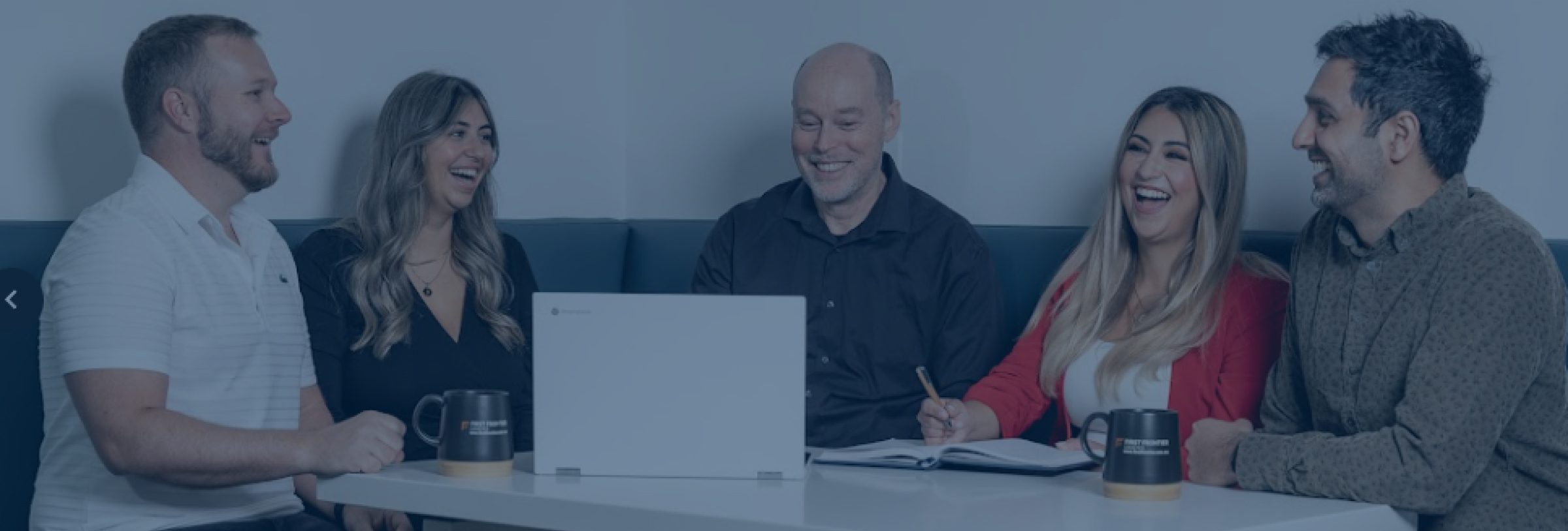 A group of five team members from FirstFrontier Logistics are seated around a table, engaged in a lively meeting, laughing and looking at a computer screen.