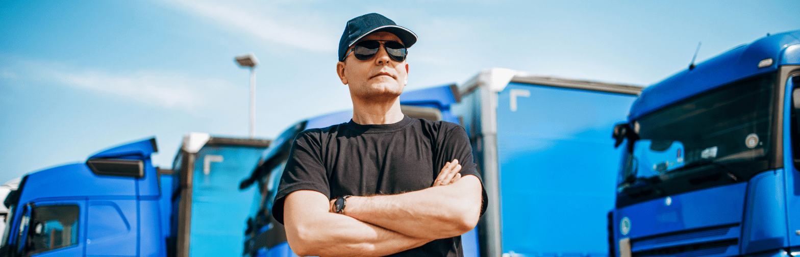 The image shows a truck driver wearing a black cap, sunglasses, and a black t-shirt, standing confidently with his arms crossed. He is positioned in front of a row of blue semi-trucks, suggesting readiness and professionalism in the transportation industry. The clear blue sky in the background adds to the vibrant and optimistic tone of the image.