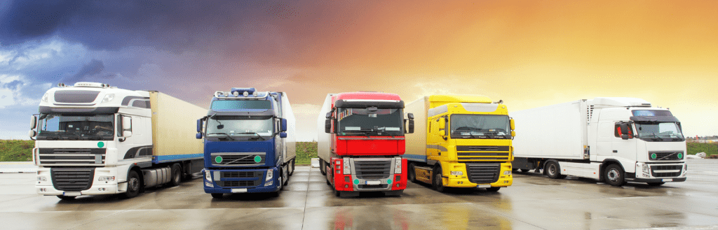 The image shows five semi-trucks parked side by side on a wet pavement with a dramatic sky in the background. The trucks are of different colors: white, blue, red, yellow, and white, arranged from left to right. The scene suggests a diverse fleet ready for Trucking Logistics, under a sky that transitions from stormy clouds to a warm sunset