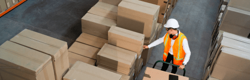 The image shows a warehouse worker wearing a white hard hat, orange safety vest, and red headphones around his neck. He is standing next to a cart filled with large cardboard boxes, surrounded by stacks of similar boxes. The worker is checking one of the boxes, indicating a busy and organized warehouse environment. Trucking Logistics