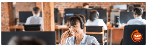Customer service representative wearing a headset and smiling while assisting a caller in a busy call center with 24/7 service, highlighting dedicated support team in a modern office environment.