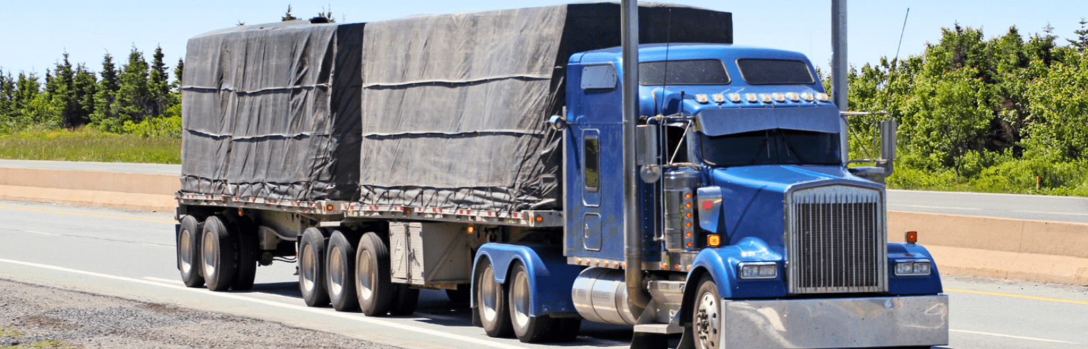 A blue semi-truck hauling a flatbed trailer covered with black tarps is traveling on a highway. The truck has multiple axles and chrome exhaust stacks, and it is driving through a scenic area with green trees and a clear sky in the background.