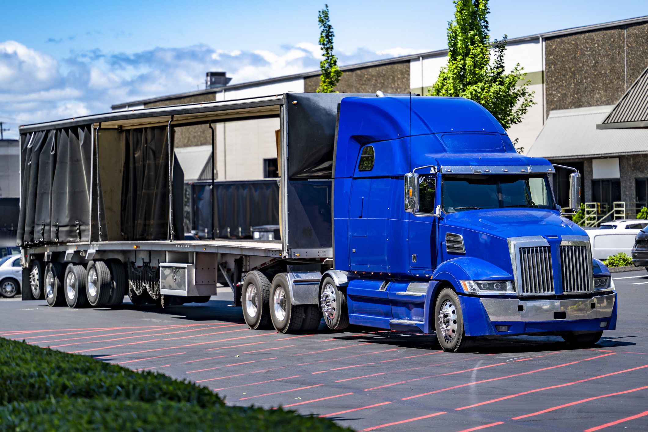 Partially open Conestoga trailer with visible side access, carrying large industrial equipment near a warehouse facility.